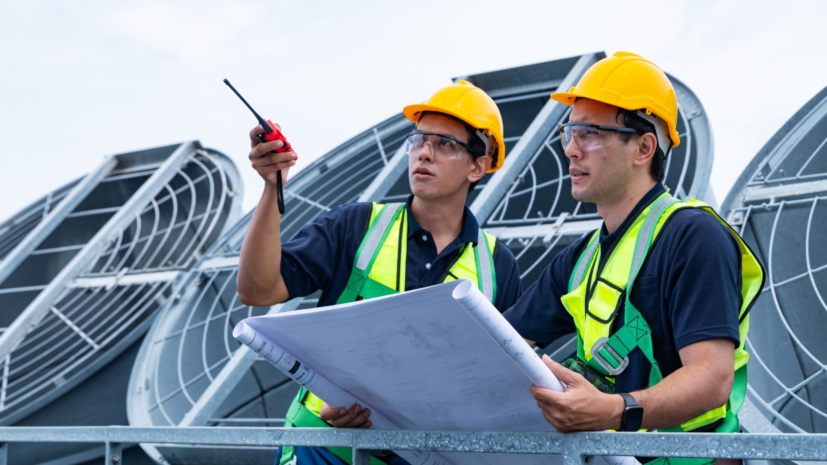 Two construction workers in safety harnesses and helmets examine technical blueprints while pointing and coordinating on a rooftop HVAC system.